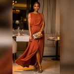 Femme adulte élégante portant une longue robe fluide terracotta, des sandales dorées et une pochette structurée dans une ambiance chaleureuse de cocktail chic.