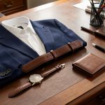 Tenue professionnelle masculine élégante avec veste bleu marine, chemise blanche, montre classique, ceinture et portefeuille en cuir, disposés avec des accessoires de bureau sur un bureau en bois.