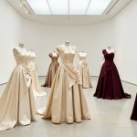 Plusieurs robes de soirée contemporaines inspirées de silhouettes historiques, présentées sur des bustes dans une galerie lumineuse, avec satin, drapés sculpturaux et volumes couture.