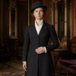 Femme adulte en redingote noire cintrée, chemise blanche et chapeau de feutre, debout dans un élégant salon victorien éclairé comme une photographie de mode.